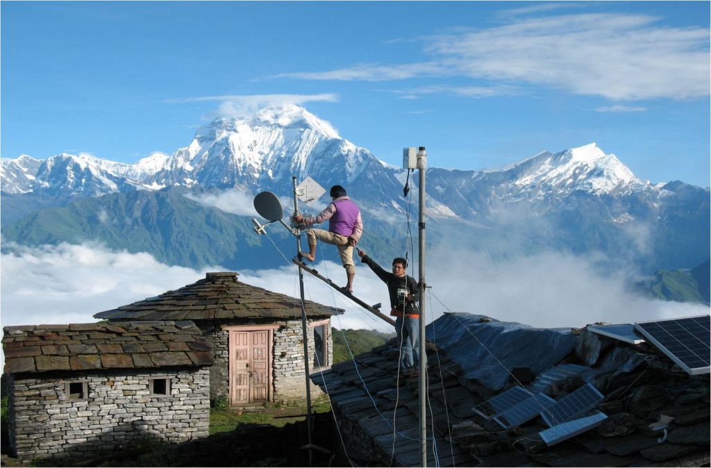 Two men fitting antennae on top of a room in front of the Himalaya mountains in Nepal