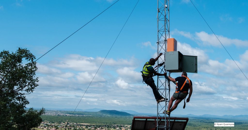 Two men work on a communications antenna in front of a blue sky and hills in the background
