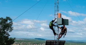 Two men work on a communications antenna in front of a blue sky and hills in the background