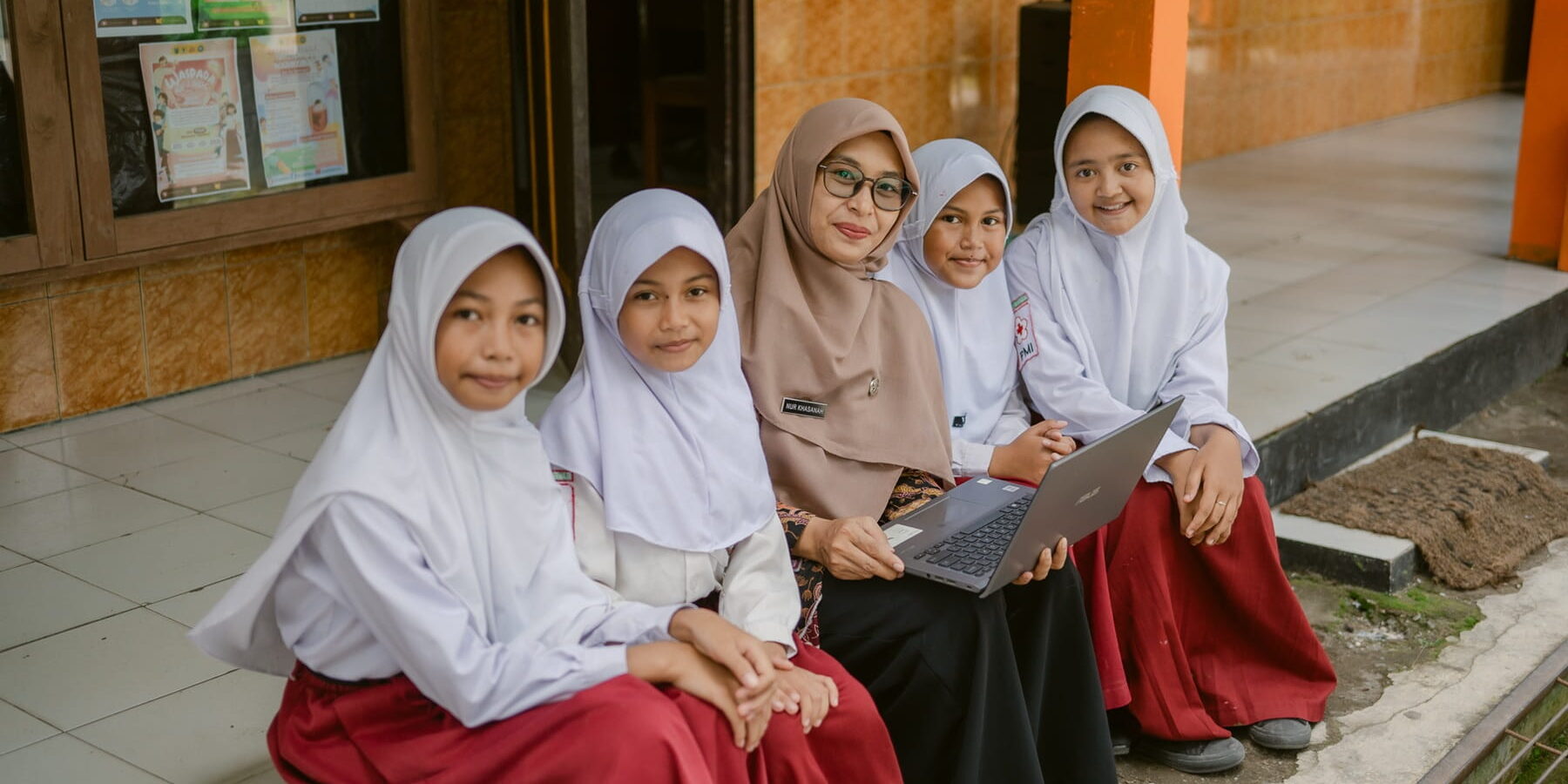 A group of girls sitting on a sidewalk with a woman in the middle holding laptop
