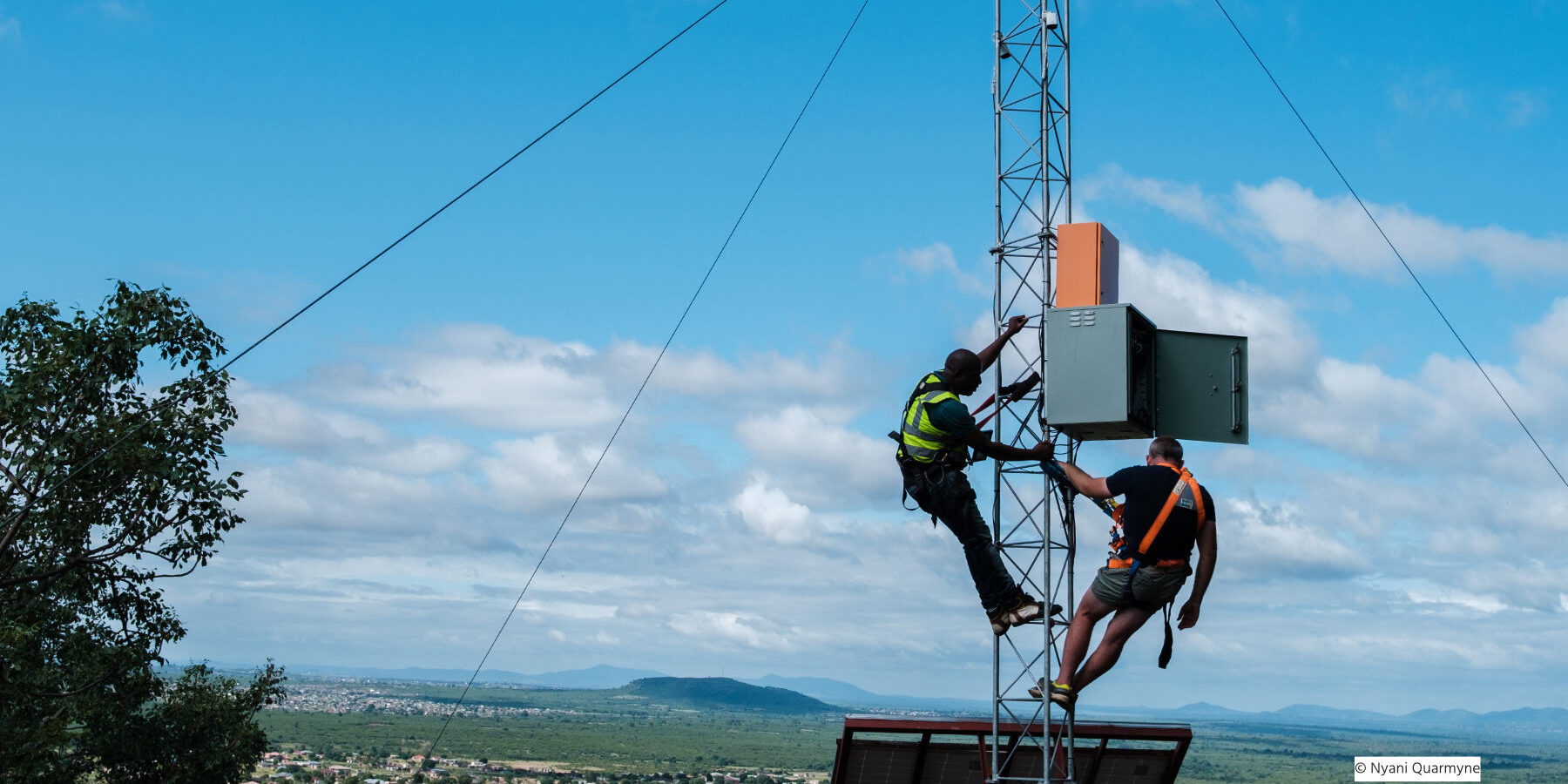 Two men work on a communications antenna in front of a blue sky and hills in the background